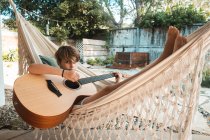 Rapaz de cabelos castanhos deitado numa rede, a tocar guitarra. — Fotografia de Stock