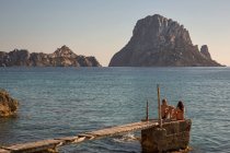 Tourist couple sitting on pier looking over to Es Vedra, Ibiza, — Stock Photo