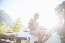 Человек, сидящий на капоте автомобиля, используя мобильный телефон — стоковое фото