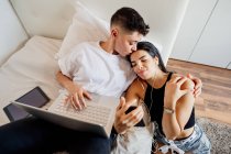Jovem casal lésbico em um quarto, usando telefone celular e laptop, beijando — Fotografia de Stock