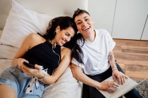 Jovem casal lésbico em um quarto, segurando telefone celular e laptop, sorrindo para a câmera — Fotografia de Stock