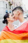 Retrato de jovem casal lésbico em pé em uma rua, envolto em bandeira do arco-íris, beijando — Fotografia de Stock
