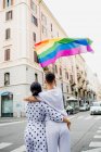 Vista trasera de una joven pareja de lesbianas de pie en una calle, ondeando la bandera del arco iris - foto de stock