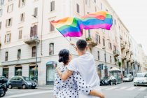 Visão traseira do jovem casal lésbico em pé em uma rua, acenando bandeira do arco-íris — Fotografia de Stock