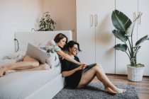 Two women with brown hair sitting on floor and lying on daybed, using laptop and digital tablet. — Stock Photo