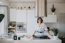 Улыбающаяся женщина с каштановыми волосами в очках, стоящая на кухне, смотрящая в камеру. — стоковое фото
