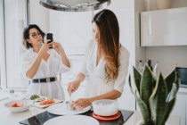 Zwei lächelnde Frauen mit braunen Haaren stehen in einer Küche, bereiten Essen zu, fotografieren mit dem Handy. — Stockfoto