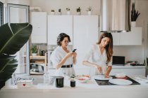 Dos mujeres sonrientes con el pelo castaño de pie en una cocina, preparando comida. - foto de stock