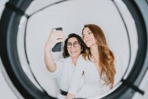 Ritratto di due donne sorridenti dai capelli castani che si abbracciano, scattando selfie con il cellulare. — Foto stock