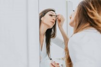 Frau mit braunen Haaren steht vor Spiegel und trägt Mascara auf. — Stockfoto