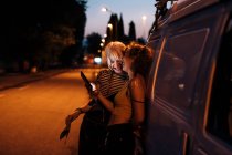 Жіноча пара сміється з телефону вночі, спираючись на фургон — стокове фото