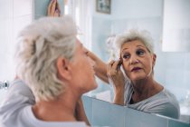 Старшая женщина смотрит в зеркало, наносит макияж — стоковое фото