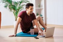 Мужчина на коврике для упражнений дома, используя ноутбук — стоковое фото