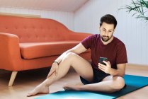 Чоловік сидить на тренувальному килимку, дивлячись на телефон — стокове фото