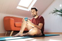 Mann sitzt auf Gymnastikmatte und schaut aufs Handy — Stockfoto