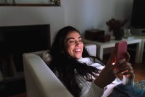 Молода жінка на дивані, посміхаючись на мобільний телефон — стокове фото