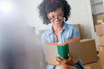 Mid adult woman reading book surrounded by cardboard boxes — Stock Photo