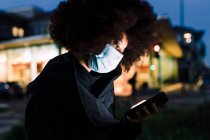 Mulher usando máscara facial, olhando para o telefone, ao ar livre à noite — Fotografia de Stock