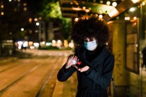 Mujer usando desinfectante de manos, usando mascarilla, al aire libre por la noche - foto de stock
