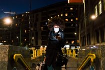 Mulher usando máscara facial, olhando para o telefone, viajando à noite — Fotografia de Stock