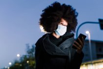 Mulher usando máscara facial e olhando para o telefone, ao ar livre à noite — Fotografia de Stock