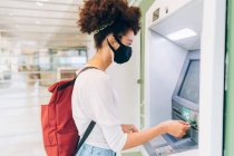 Jeune femme portant un masque facial, en utilisant ATM — Photo de stock
