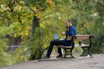 Homem no banco do parque, a atender chamadas e a trabalhar no portátil — Fotografia de Stock