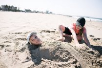 Girl burying brother in sand on beach — Stock Photo