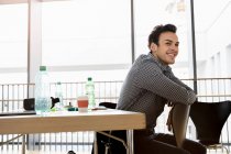 Germany, Bavaria, Munich, Young man sitting on chair by desk and smiling — Stock Photo