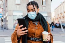 Italy, Woman in face mask holding smarphone and disposable cup on city street — стоковое фото