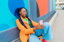 Italy, Milan, Woman with braids sitting by colorful wall, using smarphone — стоковое фото