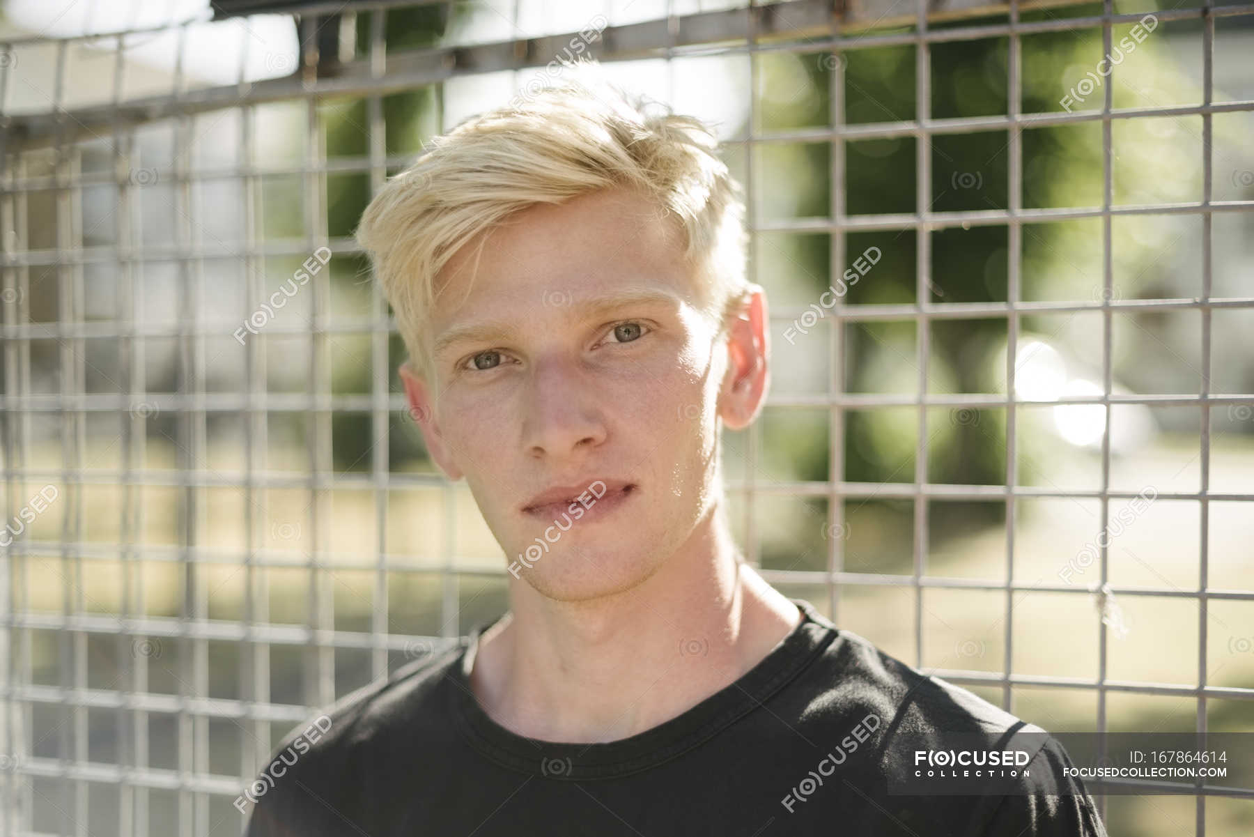 Portrait Of Blond Haired Young Man By Wire Fence Looking At
