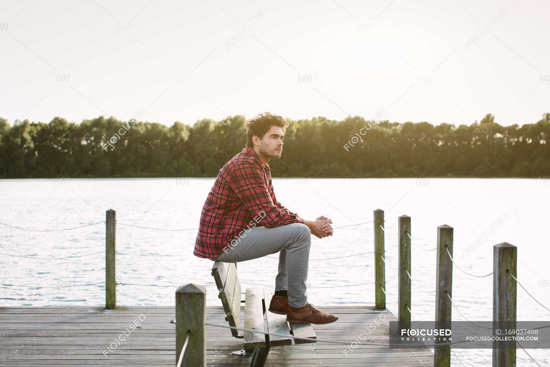 Young Man Sitting On Bench On Jetty Looking Across Water Side
