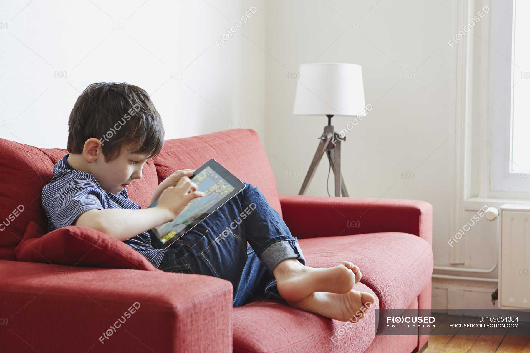 Boy using digital tablet on coach indoors — legs bent, daytime - Stock  Photo | #169054384