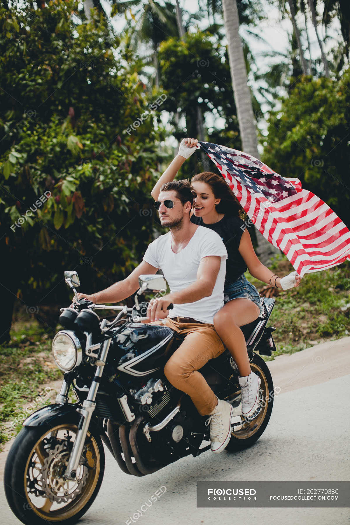 Young Couple Holding Up American Flag While Riding Motorcycle On Rural Road Krabi Thailand