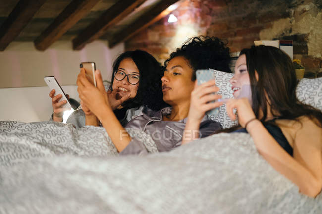 Друзья, лежащие в постели, смотрят на мобильные телефоны — стоковое фото