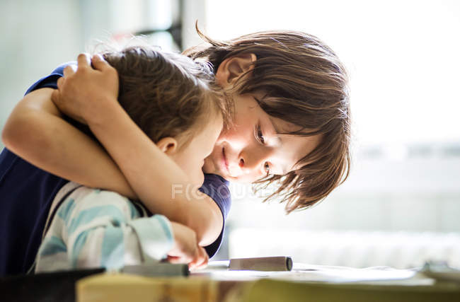 Junge am Tisch umarmt Kleinkind-Bruder — Stockfoto