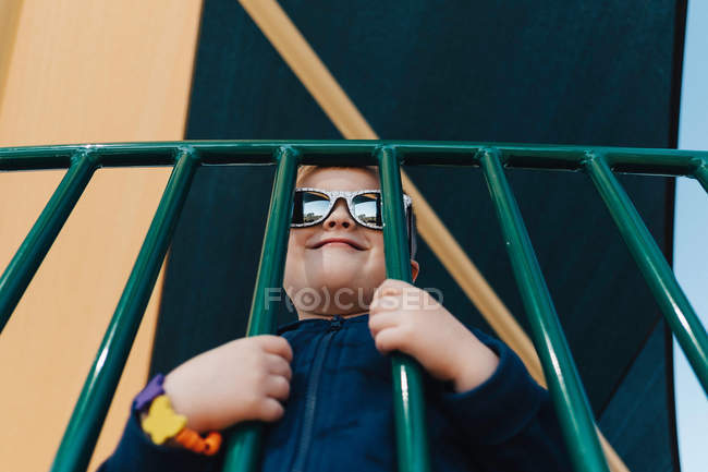 Retrato de menino usando óculos de sol olhando através de grades sorrindo — Fotografia de Stock