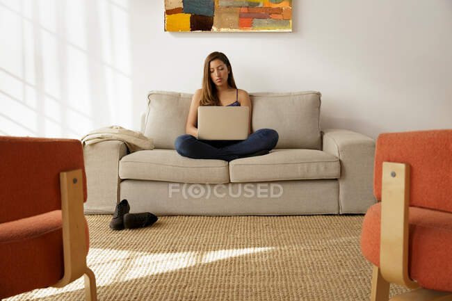 Молода жінка сидить схрещеною ногою на дивані, використовуючи ноутбук — стокове фото