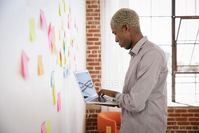 Young man standing in front of white board with sticky notes, using laptop — Stock Photo