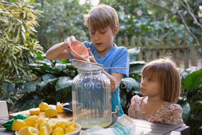 Menino e jovem irmã derramando suco de limão para limonada na mesa de jardim — Fotografia de Stock