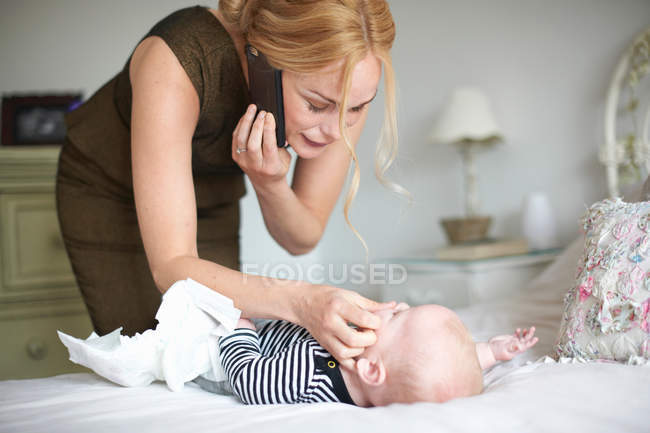 Мати розмовляє по мобільному телефону і доглядає за новонародженим хлопчиком, змінює памперс — стокове фото