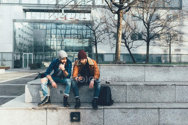 Hipster sitzen an der Wand und schauen aufs Smartphone — Stockfoto