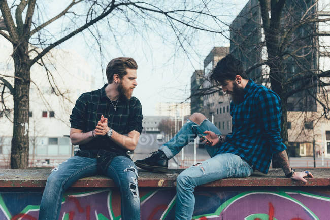 Мужчины-хипстеры сидят на стене и смотрят на смартфон — стоковое фото
