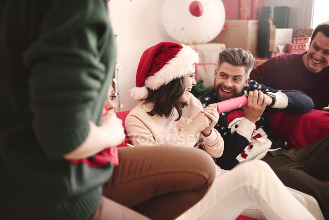 Молодые взрослые друзья тянут рождественские крекеры на диване на рождественской вечеринке — стоковое фото