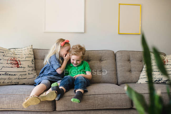 Chica en el sofá susurrando a su hermano - foto de stock