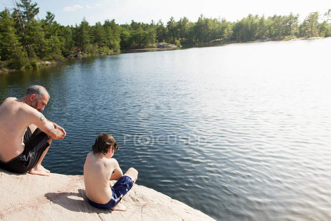 Padre e hijo sentados junto al lago quieto - foto de stock