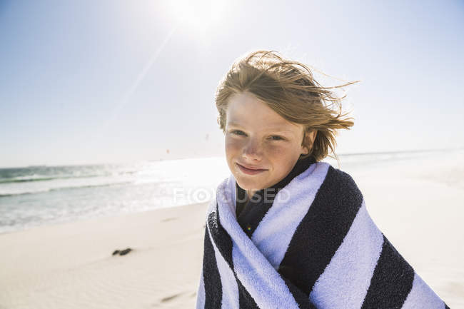 Niño en la playa envuelto en una toalla a rayas mirando a la cámara - foto de stock