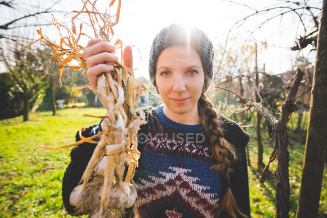 Junge Frau im Garten hält frisch gepflückte Knoblauchzwiebeln und blickt lächelnd in die Kamera — Stockfoto