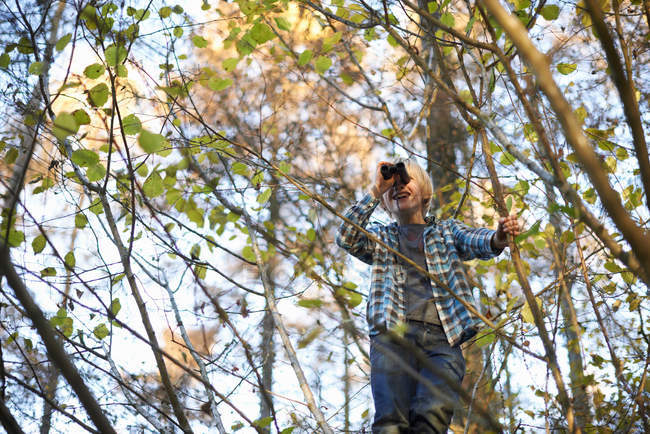 Boy on tree looking through binoculars in woods in autumn — Stock Photo